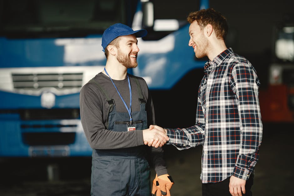Mechanic in uniform and customer in plaid shirt shaking hands in front of a blue truck inside a garage.